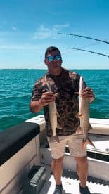 Angler proudly displaying two Speckled Trout while fishing on a sunny day in open waters.