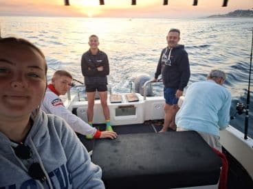 A group of four individuals enjoying a fishing trip on a boat at sunrise, with a scenic ocean backdrop.