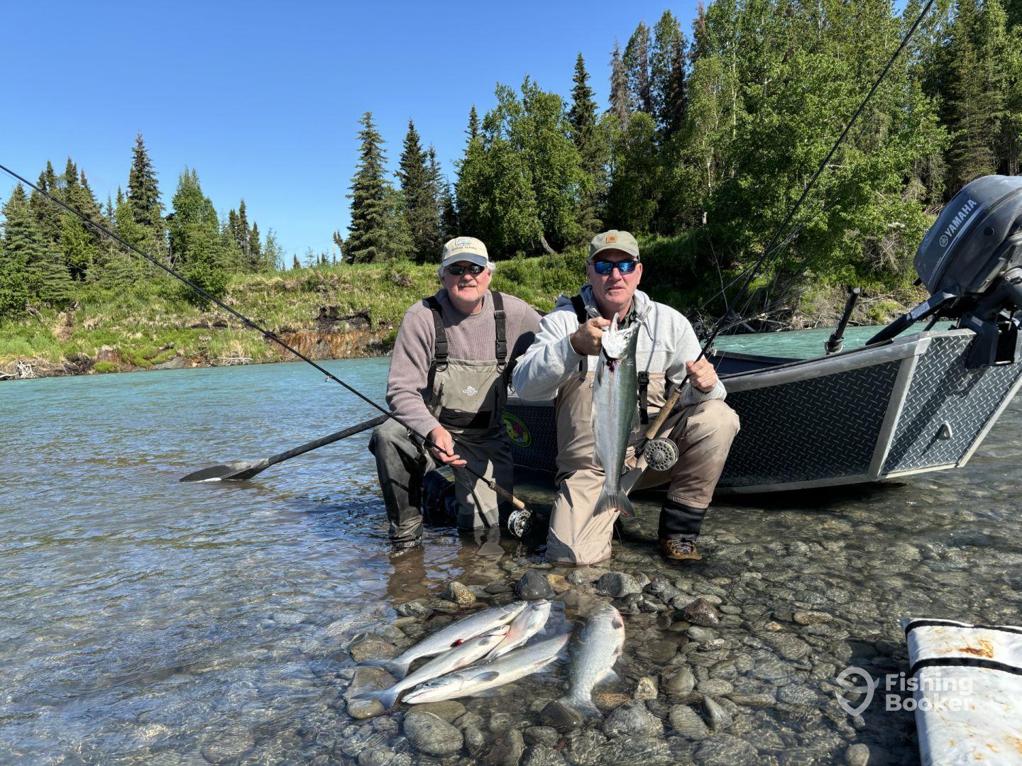 Two anglers proudly displaying their catch of silver salmon while standing in a clear river, surrounded by lush greenery.