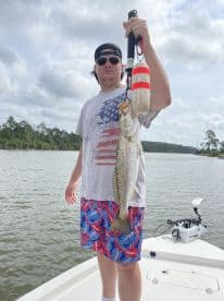 Angler displaying a large Speckled Trout while fishing from a boat in a scenic waterway.