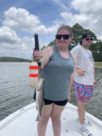 A proud angler holding a freshly caught Speckled Trout while on a boat, enjoying a day of fishing in a scenic waterway.
