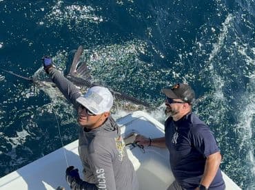 Two anglers actively reeling in a large fish, likely a billfish, while enjoying a day of deep-sea fishing on a sportfishing boat.
