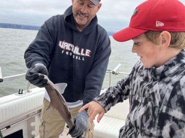 A young boy excitedly points at a small shark being held by an adult on a fishing boat, showcasing a memorable catch during their fishing trip.