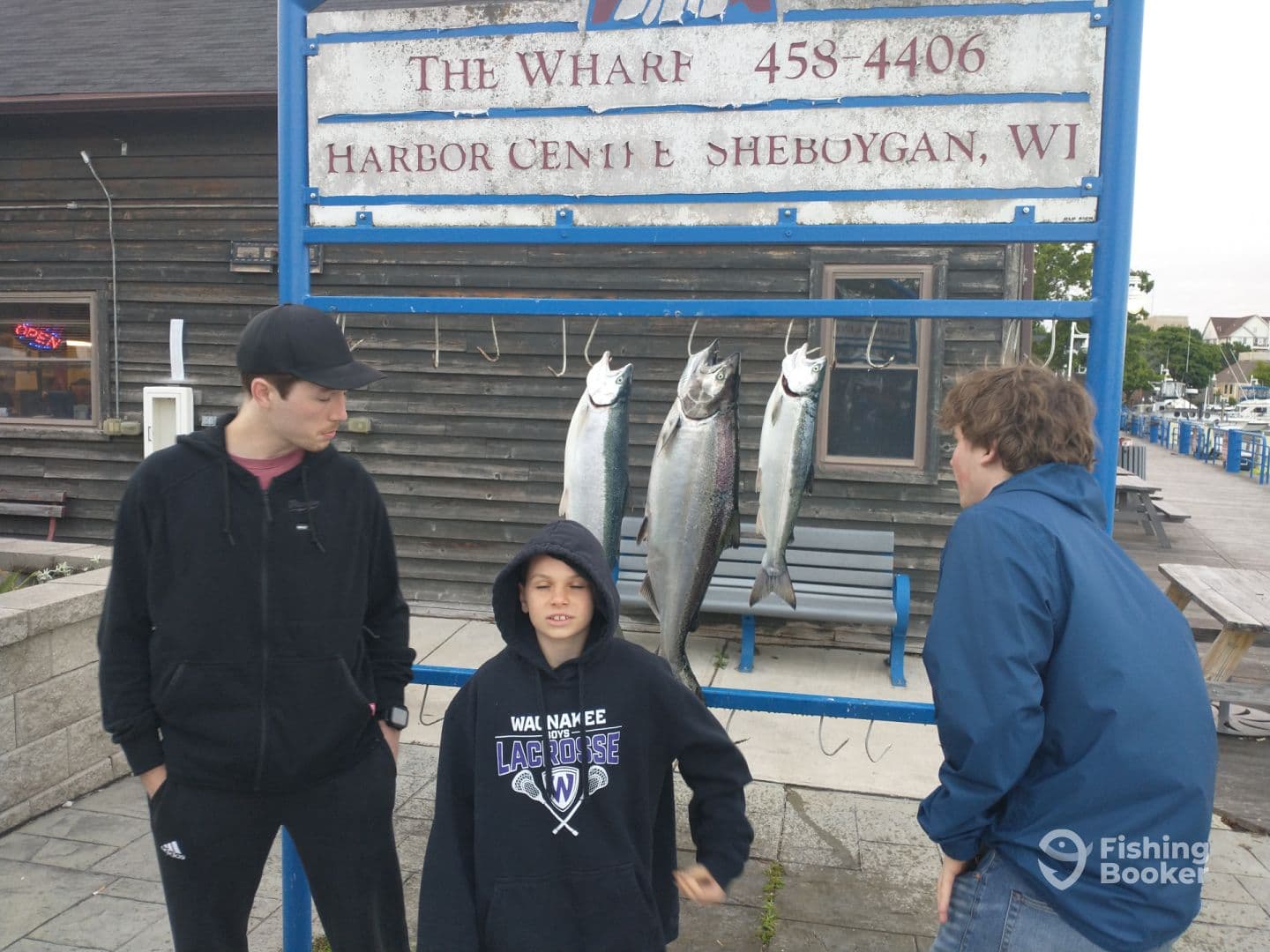 A group of three individuals, including a child, poses in front of a catch board displaying multiple fish at a Sheboygan harbor, showcasing a successful fishing trip.