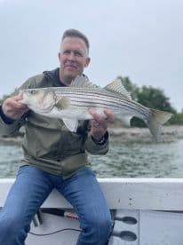 An angler proudly displaying a striped bass while fishing on a boat, showcasing a successful catch in a serene water environment.