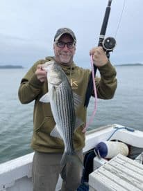 An angler proudly displaying a large Striped Bass while fishing from a boat, showcasing a successful catch in a coastal environment.