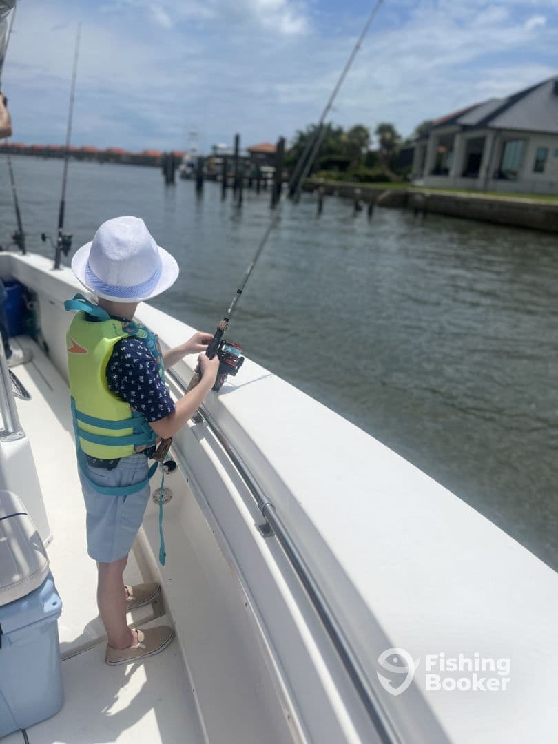 A young angler in a life jacket is casting a line from a boat, enjoying a sunny day on the water in a residential fishing area.