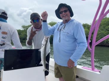 Two anglers aboard a boat, with one proudly displaying a Barracuda caught during a fishing trip in a scenic coastal environment.