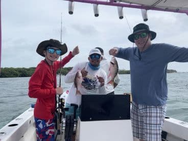 A young angler and two adults proudly displaying their catch of Snapper while fishing on a boat in a tropical setting.
