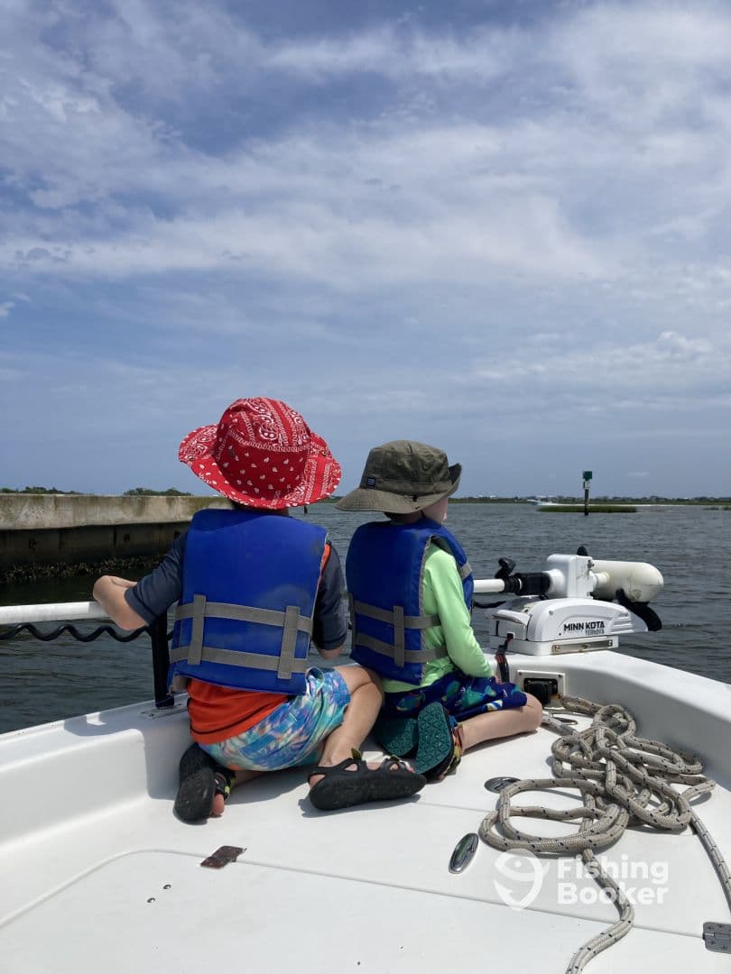 Two young boys in life jackets enjoying the view from the bow of a small boat, surrounded by calm waters and a scenic backdrop.