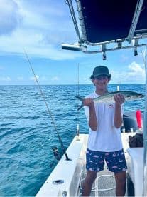A young angler proudly displaying a Spanish Mackerel aboard a fishing boat in clear blue waters.