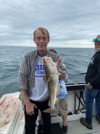 An angler proudly displaying a large Cod while fishing on a charter boat, with fellow anglers in the background.