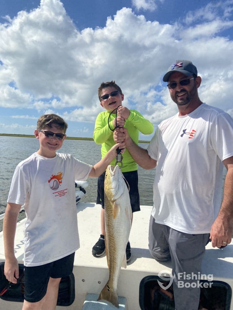 Two young anglers and an adult proudly displaying a large Redfish on a boat during a fishing trip.