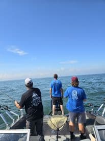 Three anglers actively fishing from a boat, casting lines into the open water under a clear blue sky.