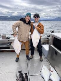 Two anglers proudly displaying their catch of halibut aboard a fishing boat in a scenic coastal environment.