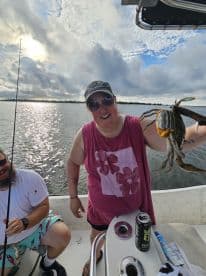 A woman joyfully displaying a large crab while on a fishing boat, with a scenic water backdrop and a fellow angler nearby.