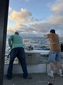 Two anglers actively fishing from the side of a boat, enjoying a day on the open water with a scenic sky backdrop.