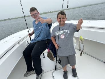 Two young anglers proudly displaying their catch aboard a boat, showcasing a flounder caught during a fishing trip.