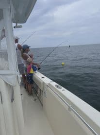 A family enjoying a fishing trip on a boat, with a child actively reeling in a fish while an adult assists. The fishing environment is calm, showcasing a cloudy sky and open water.