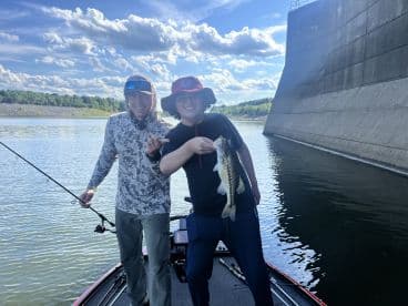 Two anglers proudly displaying a large Largemouth Bass while fishing on a lake, surrounded by scenic views and a dam structure.