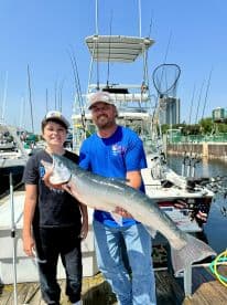 A young angler and an adult proudly displaying a large Silver Salmon at the marina, showcasing a successful fishing trip.