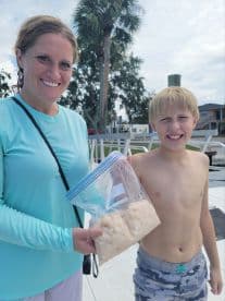 A woman and a young boy are smiling while holding a bag of scallops at a dock, showcasing their catch from a recent fishing trip.
