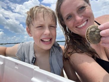 A woman and a young boy smiling while holding a scallop on a boat, enjoying a day out on the water.