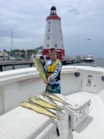 A young angler proudly displaying a large Mahi Mahi alongside several others on a boat, with a lighthouse in the background, after a successful fishing trip.