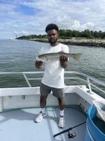 Angler proudly displaying a striped bass while on a fishing charter, with a scenic backdrop of the water and shoreline.