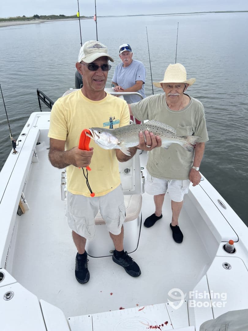 An angler proudly displaying a large Speckled Trout while fishing with friends on a boat in calm waters.