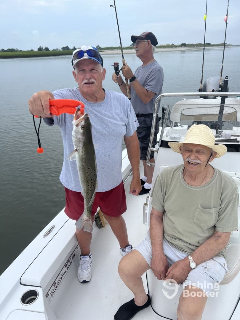 An angler proudly displaying a Speckled Trout while fishing with friends on a boat in a calm waterway.