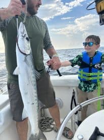 A young boy smiles while holding a large fish alongside an adult on a boat, showcasing a successful fishing trip.