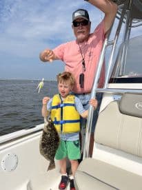 A young boy in a life jacket proudly holds a Flounder while fishing with an adult on a boat, showcasing a successful catch during a family outing.
