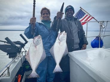 Two anglers proudly displaying their catch of Halibut while aboard a fishing boat in a coastal environment.