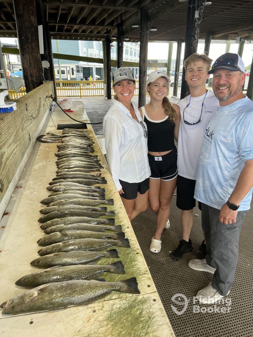 A family poses proudly in front of a catch board displaying a variety of Speckled Trout after a successful fishing trip at the dock.