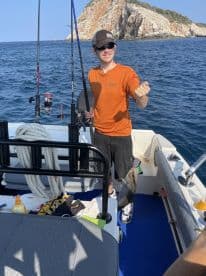A young angler reeling in a fish while enjoying a sunny day on the water, with rocky cliffs in the background.