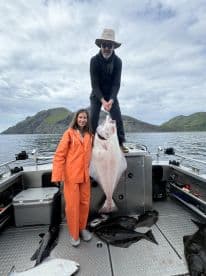 Two anglers proudly displaying a large Halibut on a fishing boat, surrounded by additional catches, in a scenic coastal environment.
