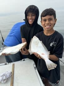 Two young anglers proudly displaying their catch of flounder while fishing on a boat.