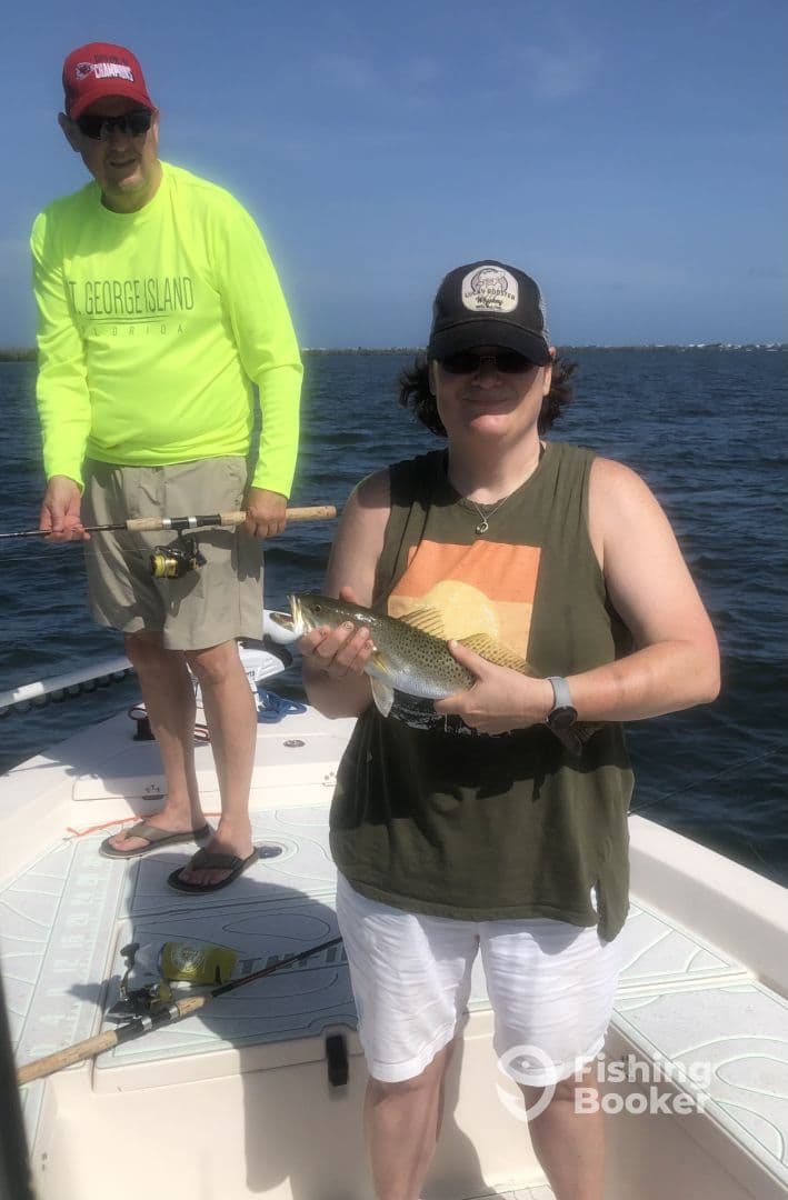 An adult angler proudly displaying a Speckled Trout while fishing on a boat in a sunny coastal environment.