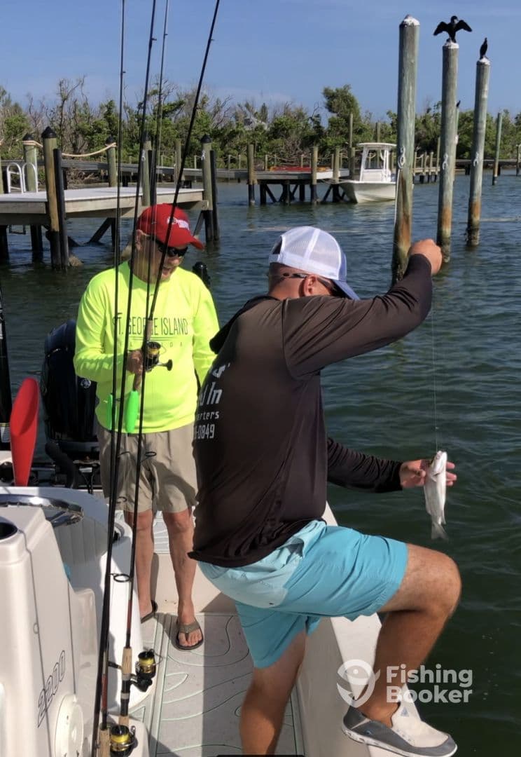 Two anglers enjoying a fishing trip, with one reeling in a fish while the other watches, set against a scenic backdrop of docks and greenery.