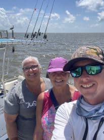 A cheerful group of three adults posing for a selfie on a fishing boat, with fishing rods visible in the background and a scenic water view.