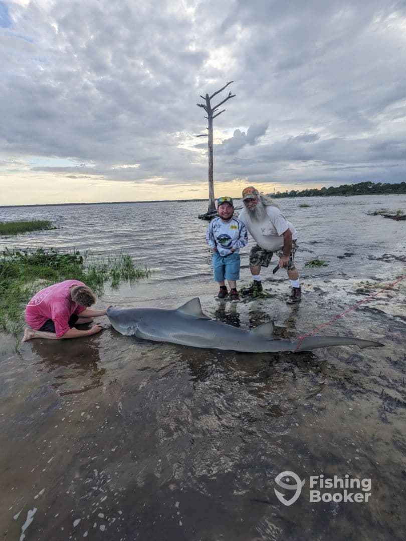 A family poses proudly with a large shark caught during a fishing trip, showcasing a beautiful waterfront setting.