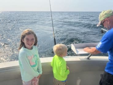Two young anglers on a boat, excitedly watching as an adult prepares a catch for display during a fishing trip.