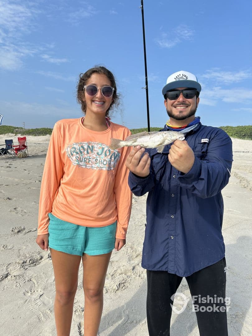 A young angler and an adult proudly displaying a caught Speckled Trout on the beach, showcasing a successful fishing outing.