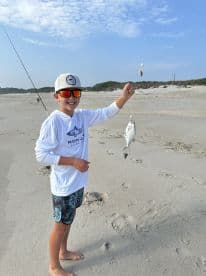 A young angler proudly displaying a caught fish on the beach, showcasing a fun day of surf fishing.