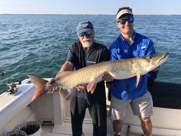 Two anglers proudly displaying a large Muskie caught during a fishing trip, showcasing the excitement of the catch against a scenic water backdrop.