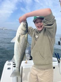A young angler proudly displaying a large Striped Bass while fishing on a boat, showcasing a joyful moment on the water.