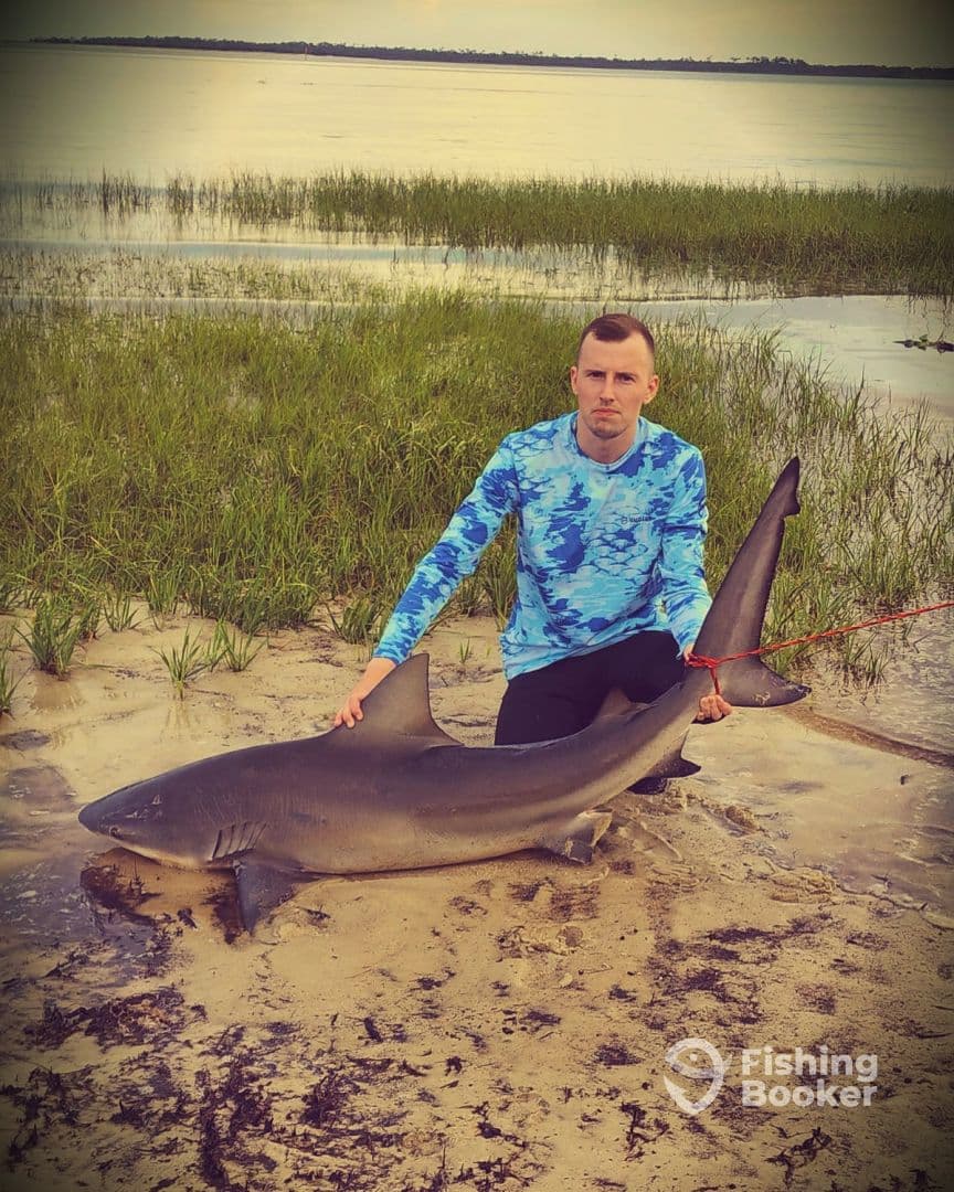 An angler proudly displaying a large shark on the sandy shore, showcasing a successful catch during a coastal fishing adventure.