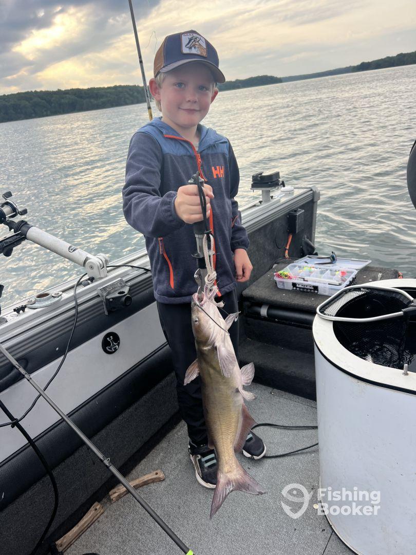 A young angler proudly displaying a Catfish while fishing on a boat, surrounded by a scenic lake backdrop.
