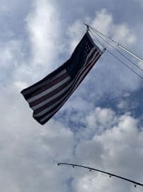 A flag waving on a fishing boat under a cloudy sky, with a fishing rod visible in the frame.